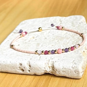 May include: A delicate bracelet featuring a light pink cord adorned with a series of small, colorful beads. The beads include pink, purple, and gold accents. The bracelet is displayed on a white, textured surface.