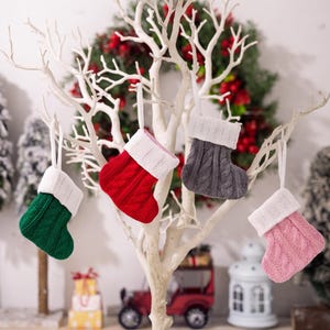 May include: Four small knitted Christmas stockings hanging from a white decorative tree. Each stocking features a cable knit pattern in a different solid color—green, red, gray, and pink—and is finished with a white ribbed cuff and a hanging loop.