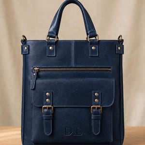 May include: A navy blue vertical tote bag featuring two top handles, a zippered front pocket, and a large flap pocket secured with two brass-toned buckles. The front flap is embossed with the letters D.L. and includes brass-toned hardware accents.