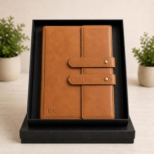 May include: A tan notebook with a smooth finish, featuring two horizontal straps secured by brass-colored metal snaps on the front cover. The initials D.L. are embossed in the bottom left corner. The notebook is presented inside a black box.