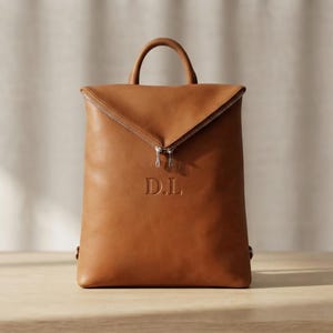 Peut inclure: Un sac à dos en cuir marron clair doté d'une poignée supérieure et d'une fermeture éclair diagonale sur la partie supérieure avant. Le devant du sac est marqué des lettres D.L. en police serif, centrées sous la ligne de fermeture.