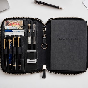 Könnte beinhalten: Ein schwarzes Leder-Organizer-Etui mit Reißverschluss und mehreren Innenfächern für Stifte, Karten und ein Notizbuch. Die rechte Seite enthält ein dunkelgraues Notizbuch mit der Prägung DUA LEATHER. Die linke Seite bietet sechs Stiftschlaufen und einen Schlüsselring.