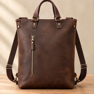 May include: A dark brown leather backpack featuring two top handles, adjustable shoulder straps, and a vertical zipped pocket on the front. The bag is embossed with the initials D.L. near the top centre and includes brass-toned metal hardware.
