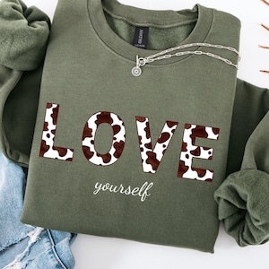 Könnte beinhalten: Ein olivgrünes Sweatshirt mit dem Wort "LOVE" in braunen und weißen Buchstaben mit Kuhmuster. Das Wort "yourself" ist darunter in Kursivschrift geschrieben. Eine silberne Halskette und blaue Jeans sind ebenfalls im Bild.