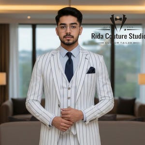 May include: A three-piece white suit with thin, vertical black stripes. The suit includes a jacket, vest, and trousers. The outfit is paired with a light blue dress shirt, a navy tie, and black dress shoes. A pocket square is visible. The text "Rida Couture Studio" is in the background.