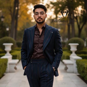 May include: A navy blue pinstripe suit with a matching jacket and trousers. The suit is paired with a brown button-down shirt. The image is taken outdoors with a blurred background of trees and greenery. The text "Rida Couture Studio" is visible.