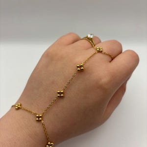 May include: A gold-colored hand chain bracelet with a delicate chain and small, clustered bead accents. The bracelet connects to a ring with a clear gemstone. The jewelry is displayed against a white background.