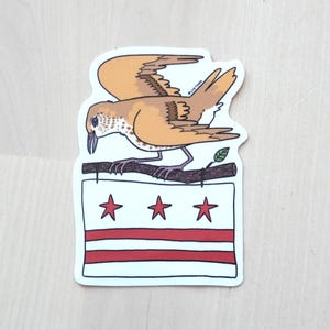 May include: A sticker featuring a cartoon bird with tan and brown feathers perched on a branch above a flag with red stars and stripes. The bird has a long beak and is facing left. The flag has three red stars and two red stripes.