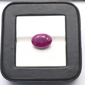 May include: A faceted oval-shaped ruby gemstone with a deep pinkish-red hue. The gemstone is displayed centered within a square black display case featuring a white interior insert that provides a high-contrast background for the stone.