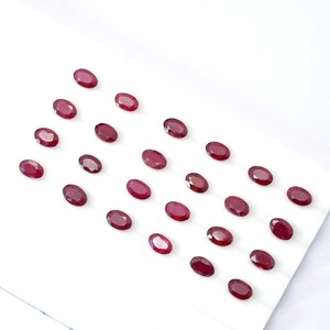 Puede incluir: Una variedad de gemas ovaladas de color rojo intenso dispuestas sobre una superficie blanca. Las gemas están espaciadas uniformemente, mostrando sus superficies pulidas y su rico color. La disposición sugiere una colección de rubíes.
