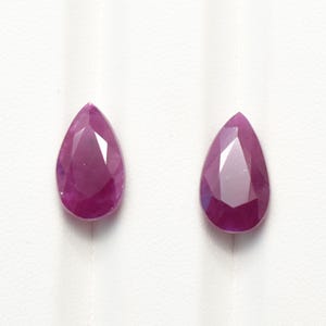 Puede incluir: Dos piedras preciosas sueltas en forma de pera con un tono rosa magenta intenso. Cada piedra presenta una superficie facetada que refleja la luz, creando un aspecto brillante y multifacético. Las piedras son simétricas y de color vibrante.