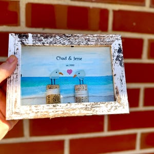May include: A framed artwork featuring two stylized seagulls perched on corks, set against a beach scene with blue and turquoise waters. The frame is white with a distressed finish. The text "Chad & Jesse est. 2002" is above the seagulls.
