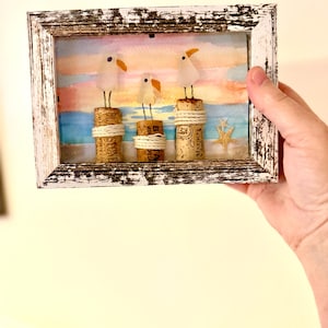 Puede incluir: Una obra de arte enmarcada con tres figuras de gaviotas posadas sobre corchos de vino, sobre un fondo de escena de playa en acuarela. El marco blanco tiene un acabado envejecido. Las gaviotas tienen picos naranjas.