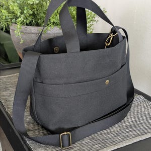 May include: A black canvas tote bag with a shoulder strap and two handles. The bag has a front pocket with a snap closure and a gold-colored metal clasp. The bag is sitting on a black table with a gray and white striped placemat.