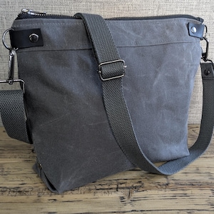 May include: A gray messenger bag with a zippered top and adjustable shoulder strap. The bag features black leather accents and silver-colored metal hardware. The bag is made of a durable, waxed canvas material.