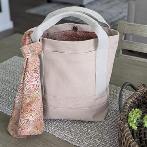 May include: A light pink tote bag with a light gray handle and a patterned scarf. The tote bag has a pocket on the front and a floral patterned interior. The bag is sitting on a gray placemat.