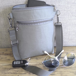 May include: A light gray crossbody bag with a zippered top and front pocket. The bag has a gray adjustable strap, a car key, and a pair of sunglasses with a silver frame. The bag is on a wooden surface.