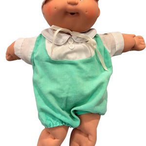 May include: A doll with a round face, dimpled cheeks, and brown eyes, wearing a white cap, a white collared shirt with two buttons, and mint green corduroy overalls. The doll has a soft, peach-toned complexion and is posed with arms outstretched.