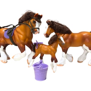 May include: Three toy horses: two adults and a foal, in shades of brown with white markings on their legs and faces. One horse has a saddle and bridle. A purple bucket with the word "WATER" is also present.