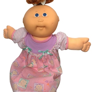 May include: A Cabbage Patch Kids doll with a round head, blue eyes, and auburn hair tied in a topknot. The doll wears a pink and purple sleeper featuring a repeating pattern of small doll faces in frames, light purple striped sleeves, and lace trim.