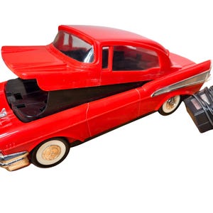Könnte beinhalten: Ein leuchtend rotes Spielzeugauto aus Kunststoff mit einem aufklappbaren Dach, das ein schwarzes Innenfach freigibt. Das Fahrzeug verfügt über silberfarbene Stoßstangen, Weißwandreifen mit goldenen Radkappen und ein schwarzes Netzteilkabel.
