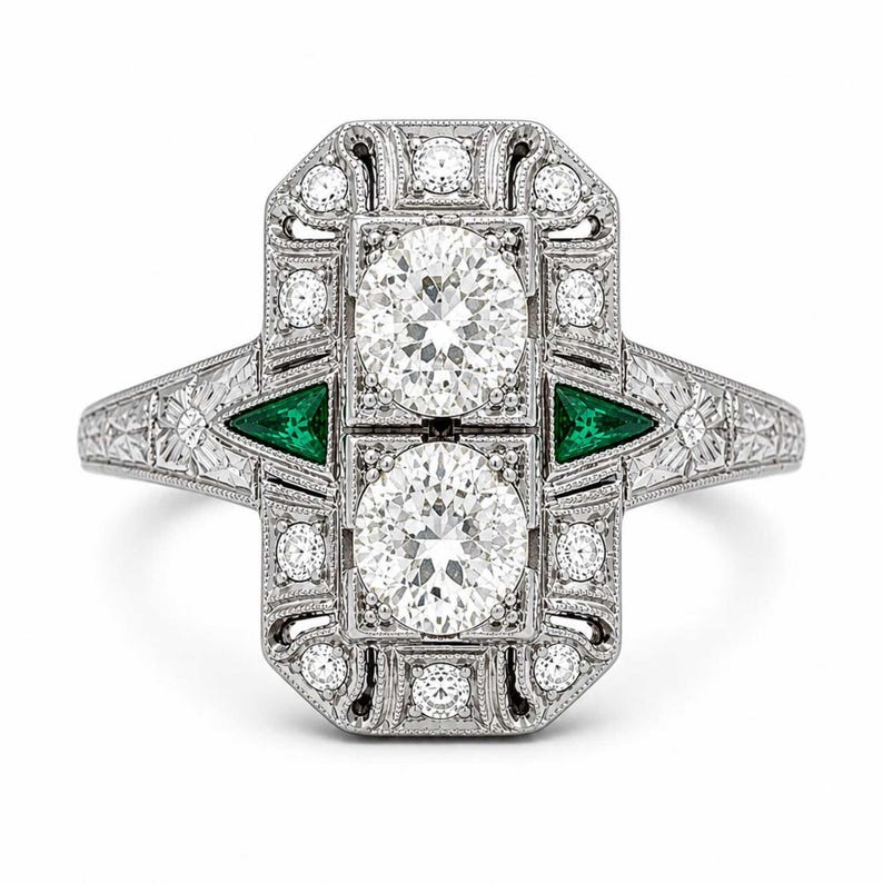 Art Deco diamond engagement ring with geometric halo design, emerald green triangular accents, and intricate vintage inspired milgrain detailing throughout.