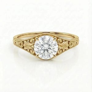 Może przedstawiać: Złoty pierścionek zaręczynowy z dużym, okrągłym, przezroczystym kamieniem szlachetnym. Opaska zdobiona jest misternymi, ozdobnymi zawijasami. Pierścionek umieszczony jest na białym tle, podkreślając jego detale i design.