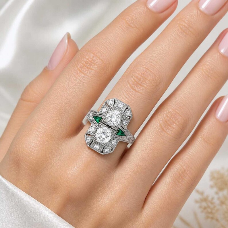 Art Deco diamond ring worn on a ring finger, showcasing the geometric halo design, sparkling diamonds, and emerald green side accents beautifully.