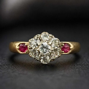 May include: A gold ring featuring a cluster of round, clear diamonds at the center, surrounded by smaller diamonds. Two round, red rubies are set on either side of the ring's band. The ring is set against a dark background.
