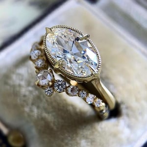May include: A gold engagement ring set featuring a large, oval-cut diamond with a halo setting. The band is adorned with smaller diamonds. The ring is set against a soft, neutral background, highlighting the sparkle of the stones.