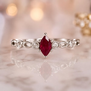 May include: A silver-toned ring featuring a central marquise-cut deep red ruby gemstone flanked by two small round white diamonds on each side, set within a delicate metal band with an intricate openwork pattern of repeating oval shapes.