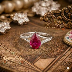 May include: A silver ring with a pear-shaped, deep red gemstone as the focal point. The ring's band splits into two, with small, clear stones set along the split. The ring is displayed on a textured, brown surface.