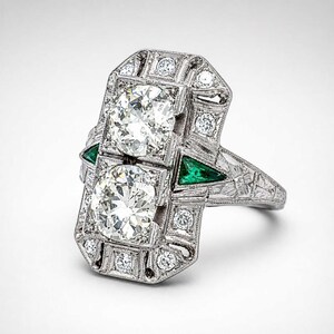 Antique style diamond statement ring featuring two round brilliant center stones surrounded by sparkling accents and vivid green side gemstones.