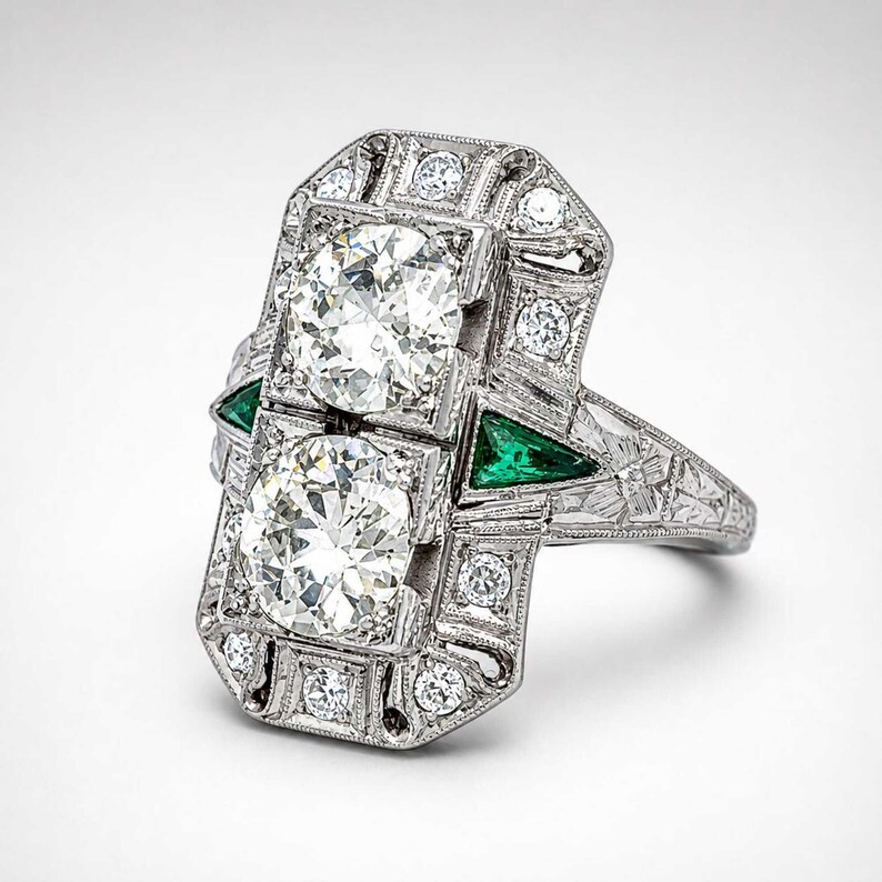 Antique style diamond statement ring featuring two round brilliant center stones surrounded by sparkling accents and vivid green side gemstones.