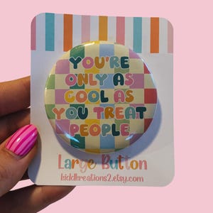Könnte beinhalten: Ein großer, farbenfroher Button mit dem Text "YOU'RE ONLY AS COOL AS YOU TREAT PEOPLE" in einer Retro-Schriftart. Der Button befindet sich auf einer weißen Karte mit Streifenmuster und den Worten "Large Button".