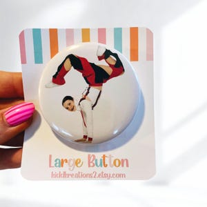 May include: A large button featuring a dancer in a handstand, wearing a red, black, and white costume. The button is on a white card with a colorful striped border. The text "Large Button" and a website address are printed on the card.