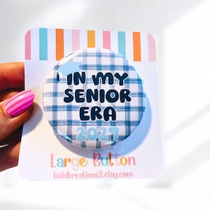 Könnte beinhalten: Ein großer, runder Button mit dem Text "IN MY SENIOR ERA 2021" in Marineblau, vor einem blau-weiß karierten Muster. Eine Grafik einer Abschlusskappe befindet sich über dem Text. Der Button befindet sich auf einer weißen Karte mit bunten Streifen.