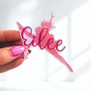 May include: A pink sticker featuring a silhouette of a person pole dancing with the name "Eilee" in a cursive font. The sticker has a glittery texture and a white outline.