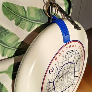 Puede incluir: Disco volador blanco de Bolonia con un gráfico de mapa y detalles en azul. El disco tiene un asa azul con la palabra "Poppi" impresa. El disco está sujeto a un cordón negro y un clip de metal plateado.