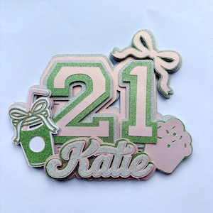 Op de afbeelding: Een gelaagde taarttopper met het getal 21 in groen en lichtroze glitterkarton. De naam Katie staat in zilverkleurige glittercursief onder het getal. Decoratieve elementen zijn een groene glitterdrankbeker en een roze cupcake.