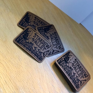 Op de afbeelding: Verschillende rechthoekige bruine leren patches, elk met de woorden "Stardew Valley" in het zwart. De patches hebben een zwarte illustratie van een landschap met gebouwen en bomen.