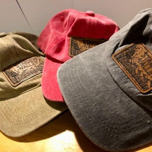 Puede incluir: Tres gorras de béisbol en verde oliva, rojo y gris. Cada gorra tiene un parche de cuero con las palabras "Stardew Valley" y una escena de montaña. Las gorras están hechas de una tela suave y desgastada.