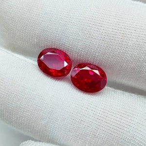 Puede incluir: Dos gemas rojas vibrantes de corte ovalado. Las gemas se muestran sobre una tela blanca texturizada. Las piedras tienen un alto brillo, reflejando la luz. Estas gemas son probablemente rubíes, a menudo utilizados en joyería.