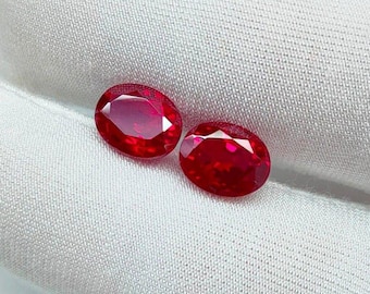 May include: Two oval-cut, vibrant red gemstones. The gems are displayed on a white, textured fabric. The stones have a high luster, reflecting light. These gemstones are likely rubies, often used in jewelry.