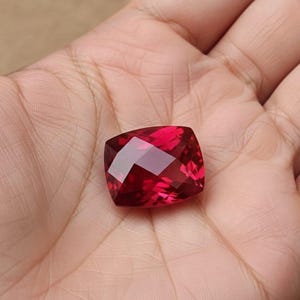 May include: A faceted, square-cut, deep red gemstone. The gem is held in a hand, showcasing its brilliant, reflective surfaces. The stone's color is a rich, saturated red, with multiple facets catching the light.