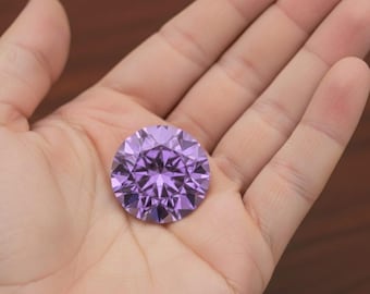 Könnte beinhalten: Ein großer, runder, facettierter lila Edelstein, der in einer offenen Hand gehalten wird. Der Edelstein hat einen Brillantschliff, der das Licht reflektiert. Die Farbe ist ein lebendiger Violettton. Der Hintergrund ist eine verschwommene Holzoberfläche.