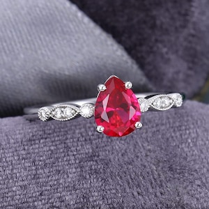Pode incluir: Um anel de prata com uma pedra preciosa vermelha vibrante em forma de pêra como ponto focal. A banda é adornada com pequenas pedras claras e detalhes decorativos. O anel é apresentado sobre um fundo cinza suave.