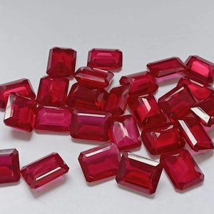 May include: A collection of radiant, rectangular-cut rubies. The gemstones are a deep, saturated red, with facets that catch the light. The rubies are scattered across a white surface, showcasing their vibrant color and clarity.