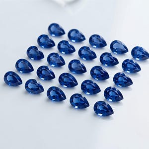 May include: A collection of twenty-four loose, pear-shaped faceted gemstones in a deep, vibrant royal blue color. Each stone features a pointed top and a rounded base, arranged in a grid pattern on a plain, light-colored surface with clear facets.