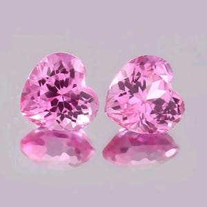 May include: Two heart-shaped, faceted pink gemstones. The gems are a vibrant pink color and reflect light, creating a sparkling effect. The gemstones are displayed on a reflective surface, enhancing their brilliance.
